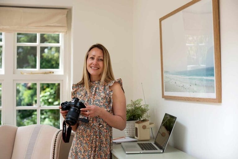 An image of a woman stood in a room holding a camera. She has long blonde brown straight hair and smiles to camera. She wears a long floral dress and is stood next to a dresser with an open laptop on it.