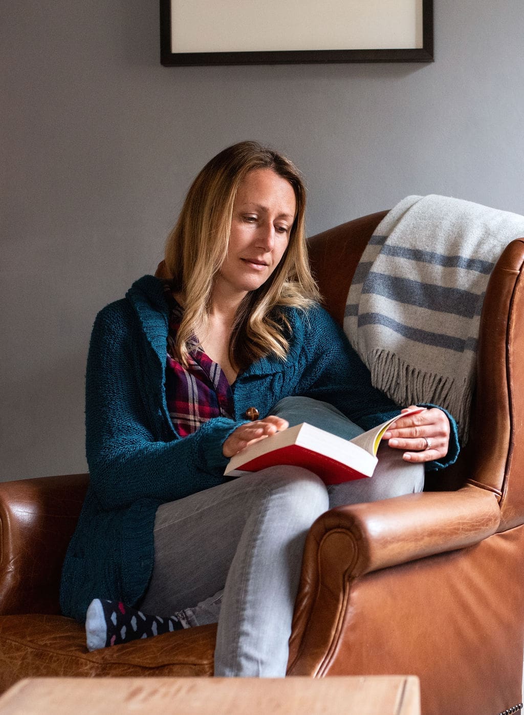 An image of a woman sat in a brown leather armchair reading a book. She has long blonde brown hair and wears a teal cardigan over a red checked shirt and light grey jeans. A folded blanket with grey stripes is hung over the back of the chair.