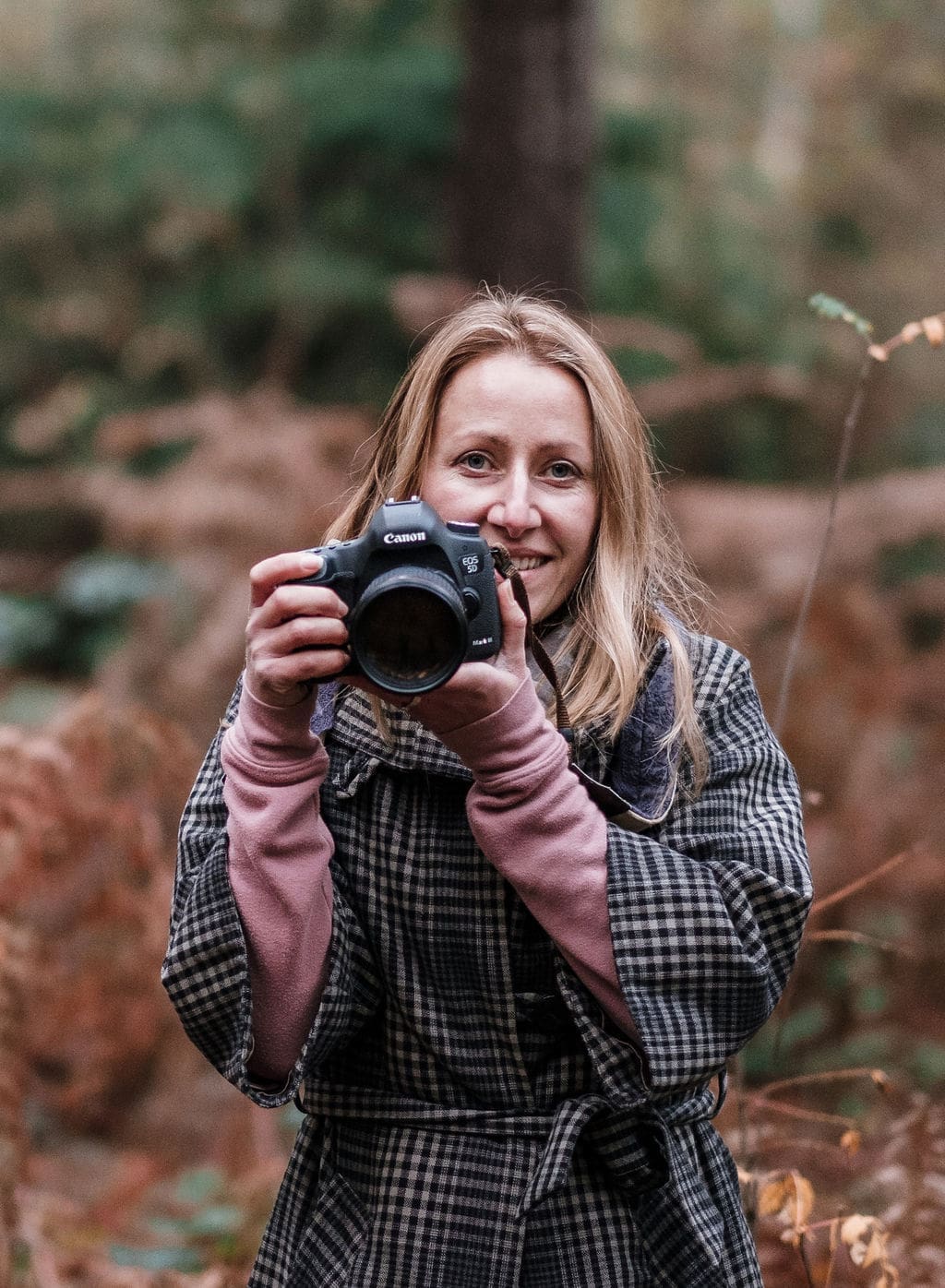 An image of a woman in an autumnal setting. She has long blonde brown straight hair and smiles from behind a camera. She wears a dark checked jacket over a light pink jumper.