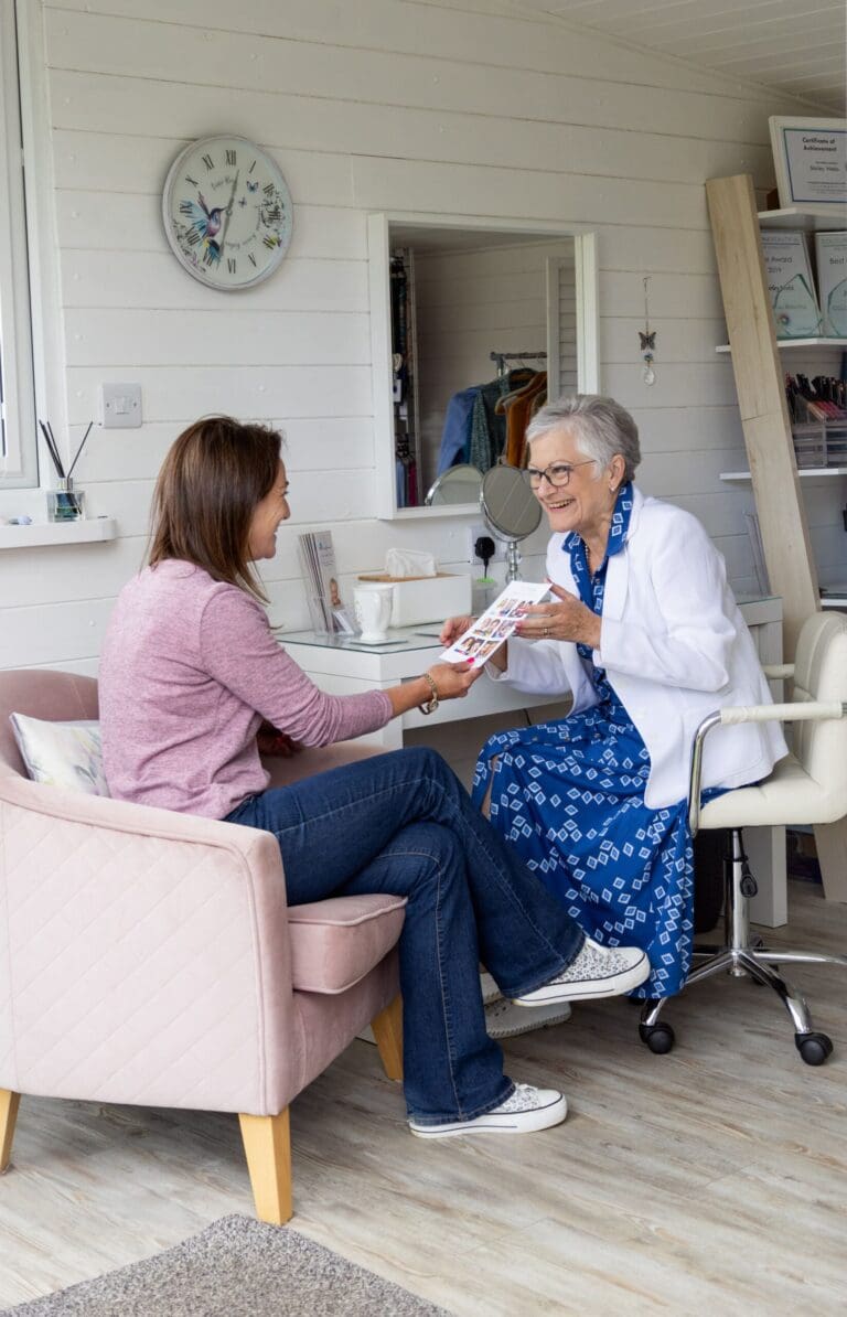Two women sit facing each other looking at a document. The lady on the right has short grey hair and wears glasses, a blue patterned dress and a white jacket. She's smiling at the other lady who has long dark hair and weras a duky pink jumper over blue jeans.