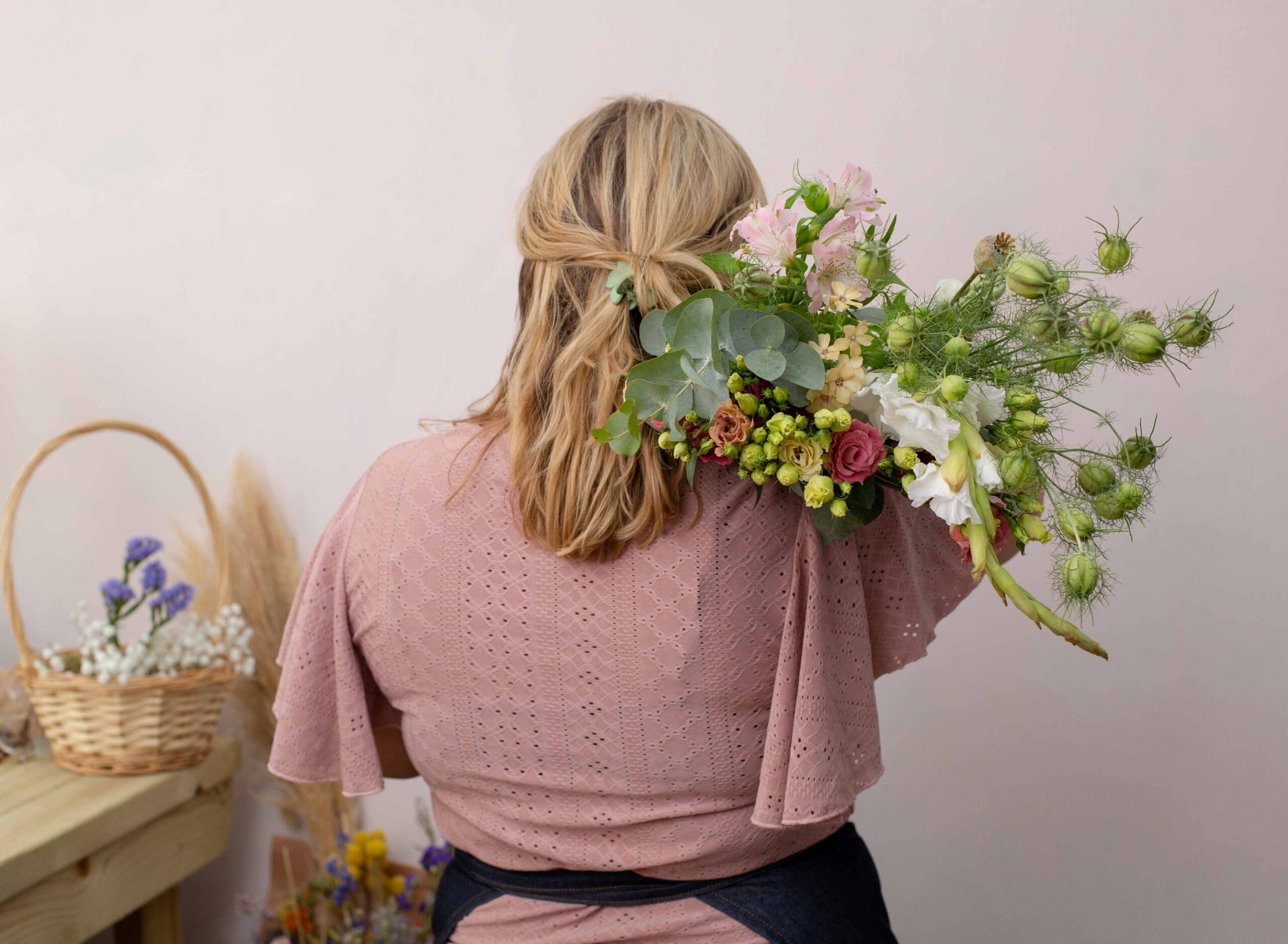 An image of a woman photographed from behind, holding a bouquet of flowers over her shoulder. She wears a pale pink top and a navy apron is tied around her waist.