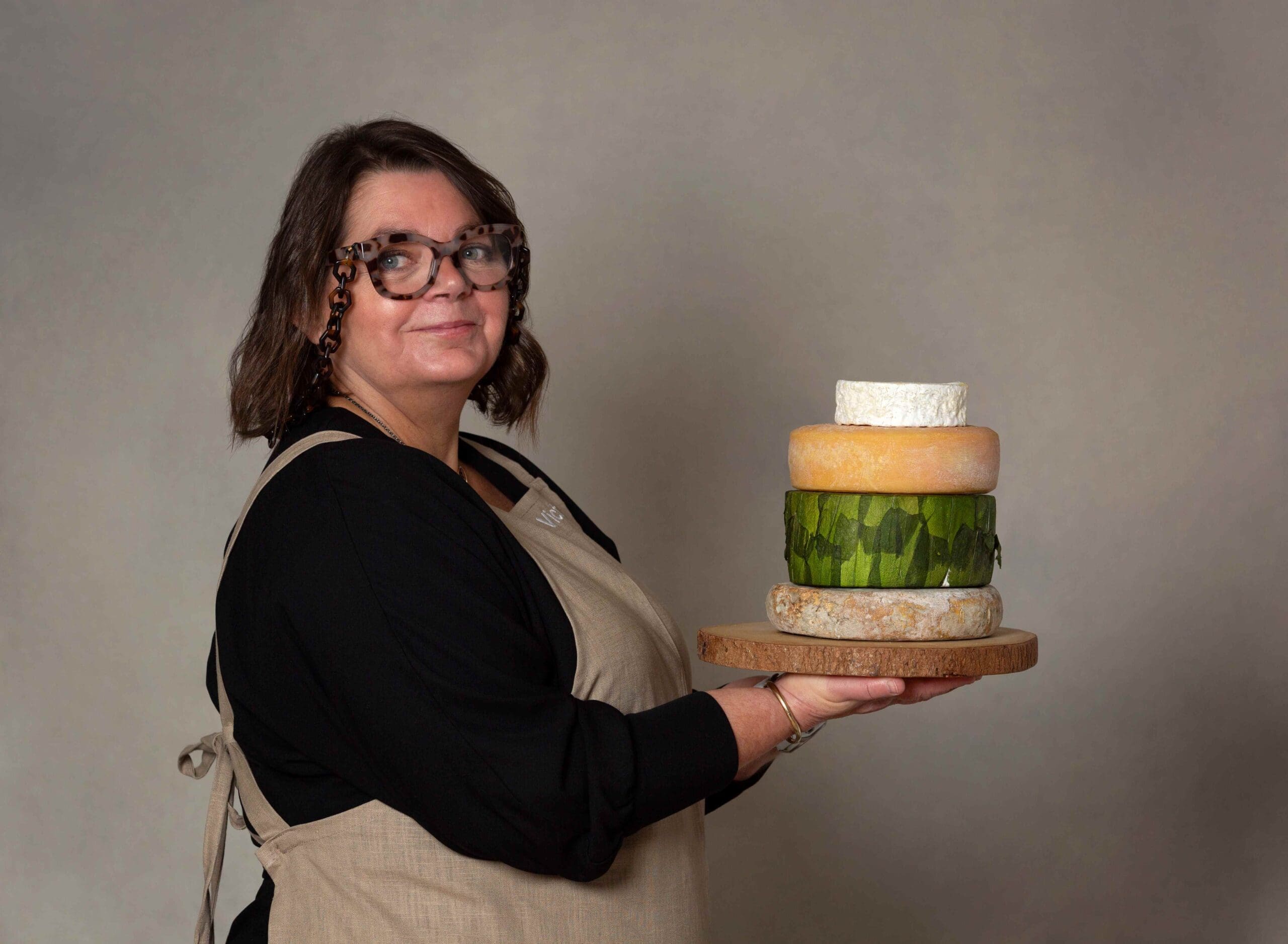A woman photographed in a studio against a grey beige backdrop. She's stood side on and holds a wooden board in front of her with a stack of 4 cheeses on top. She has shoulder length dark borwn hair and wears a black shirt and beige apron.