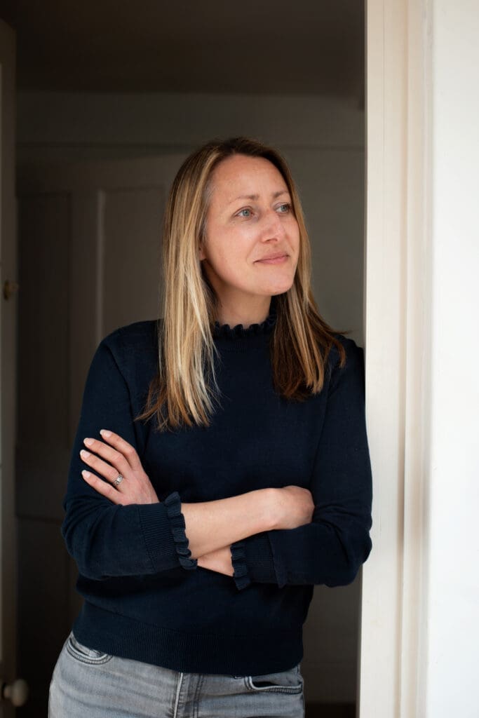 A woman with long brown blonde hair leans on a doorframe indoors. She wears a dark jumper with grey jeans and is looking off to the side with a slight thoughful smile.
