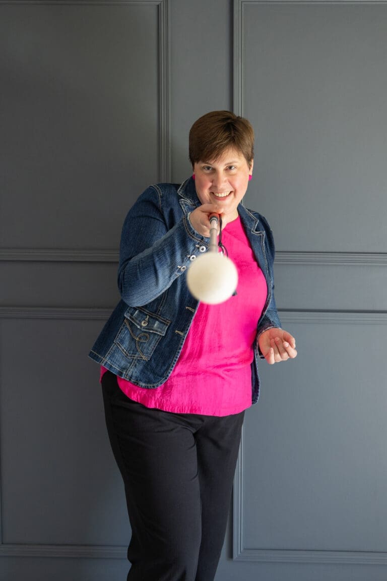 A woman photographed against a grey pannelled wall. She is holding out a white cane in front of her pointed directly at the camera, the end of which is going out of focus. She's looking along the cane at the camera. She wears a bright pink top under a denim jacket with black trousers. She has a big grin.