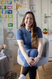 A young woman with long dark wavy hair sits in a chair smiling to camera. She wears a long blue, short sleeve dress and has her arms reaching forward holding her knee. She's indoors with lots of posters on the wall behind her.