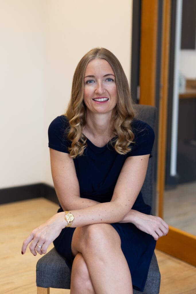 An image of a woman with long wavy blonde-brown hair. She sits in a chair and leans towards camera with her arms loosely crossed on her knee. She's wearing a navy dress.