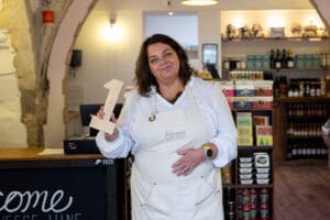 A cheesemonger stood in her shop holding a wooden number one. She has shoulder length dark hair and smiles to camera. She's wearing a cream apron over a white shirt. 