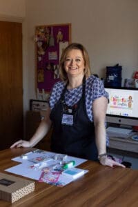 An image of a woman with shoulder length hair standing at a table. She's wearing a black apron over a patterned short sleeve shirt and smiles to camera. In front of her on the table is a craft kit laid out. 